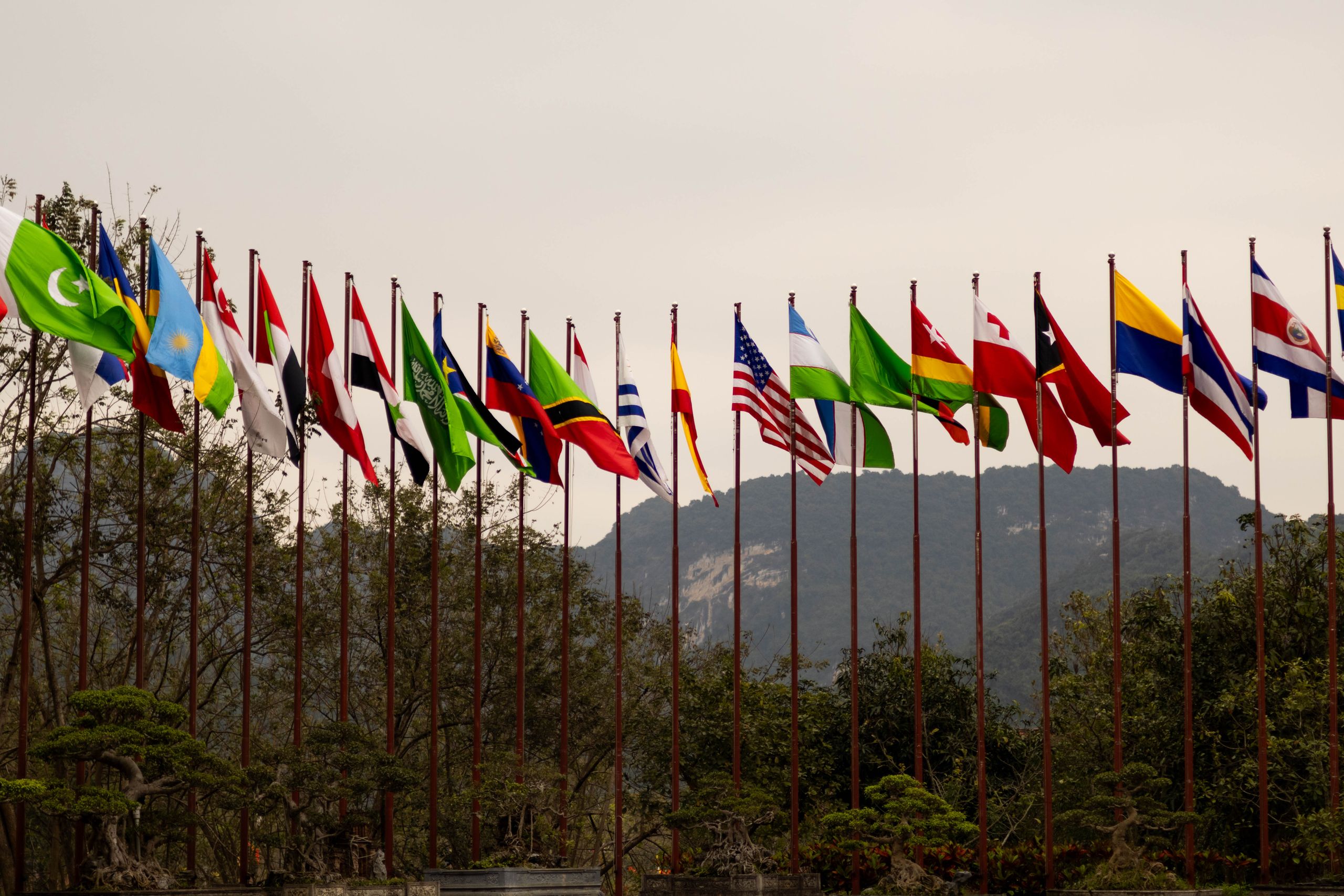 A display of various national flags on flagpoles set against a mountain backdrop, symbolizing global unity.